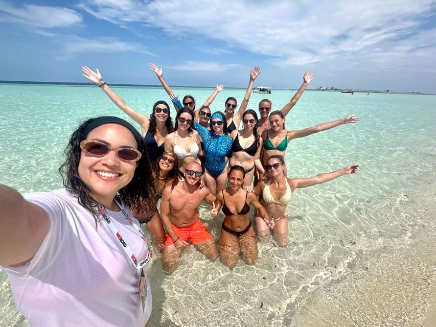 Un groupe WeRoad souriant pour un selfie, les pieds dans l'eau turquoise peu profonde et claire.