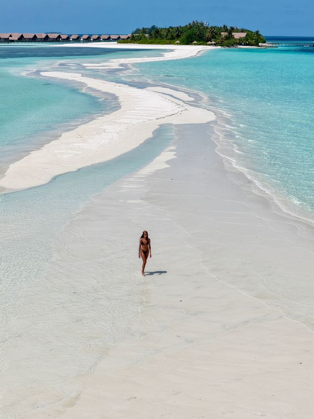 Une femme en bikini marche le long d'un banc de sable blanc sinueux, entourée d'eaux turquoise claires, avec une île tropicale en arrière-plan.