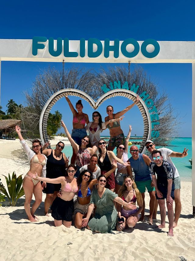 Un groupe WeRoad posant pour une photo sur une plage de sable blanc devant un cadre en forme de cœur sous un ciel ensoleillé.