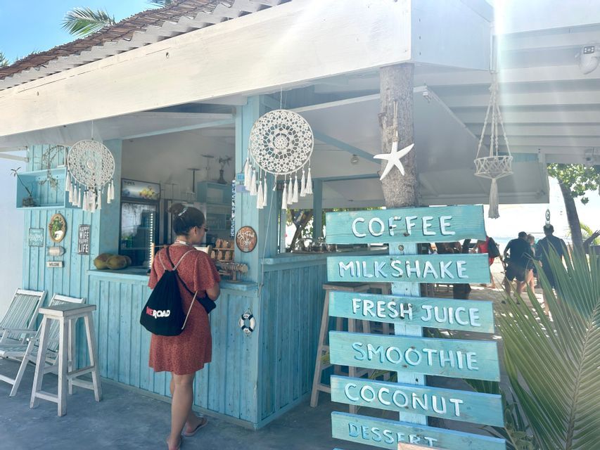 Une personne avec un sac à dos WeRoad commande au comptoir d'un kiosque de plage en bois bleu clair, par une journée ensoleillée.