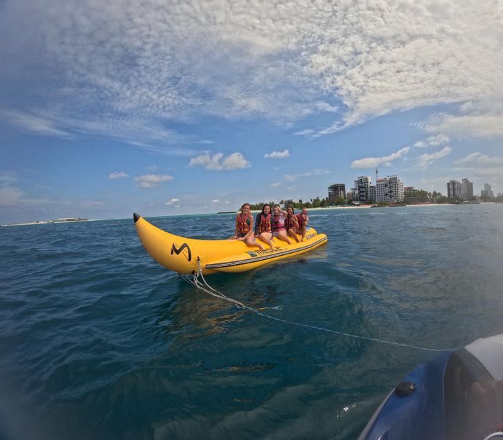 Un groupe WeRoad de cinq femmes en gilets de sauvetage sur un bateau banane jaune gonflable en mer, avec une ville à l'horizon.