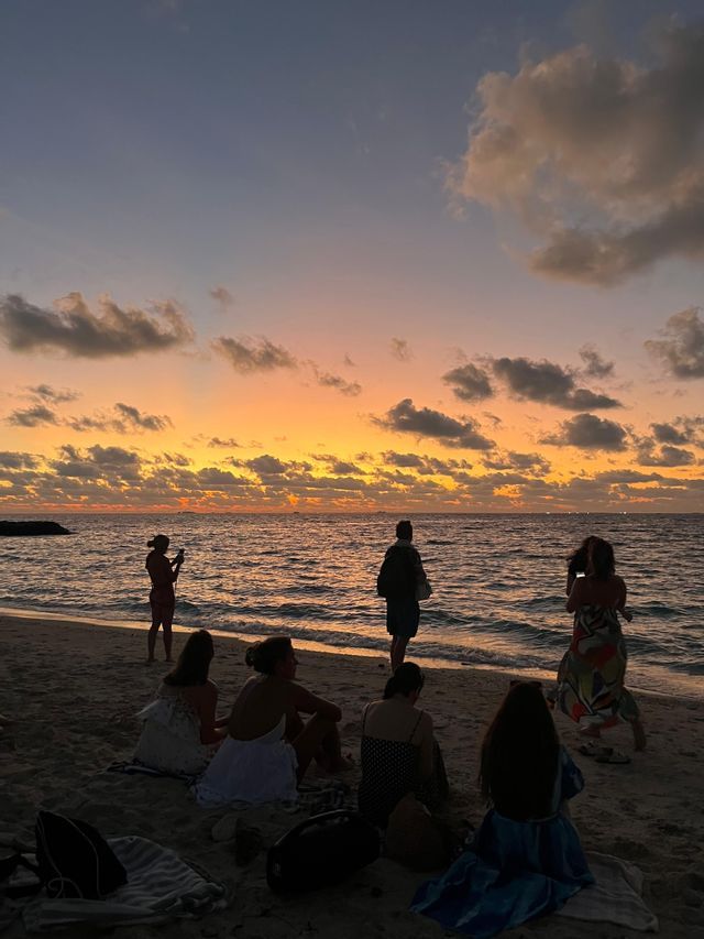 Un groupe WeRoad en silhouette est assis et debout sur une plage, admirant un coucher de soleil coloré sur l'océan.