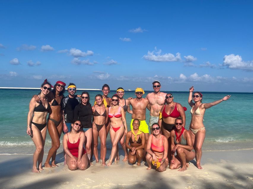 Un groupe WeRoad en maillot de bain posant pour une photo sur une plage de sable au bord de l'océan turquoise.