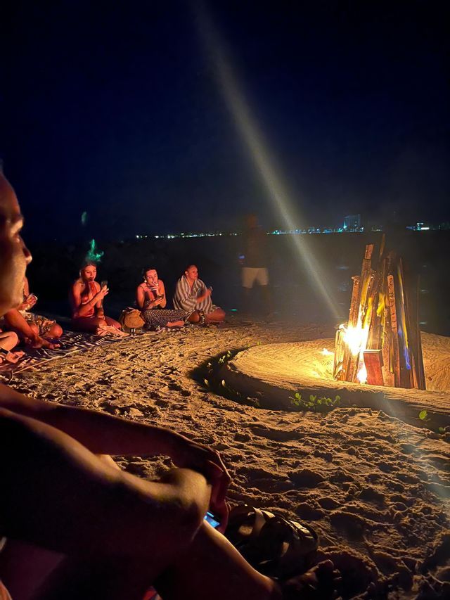 Un groupe WeRoad est assis en cercle autour d'un feu de joie sur une plage de sable la nuit, avec la skyline d'une ville en arrière-plan.