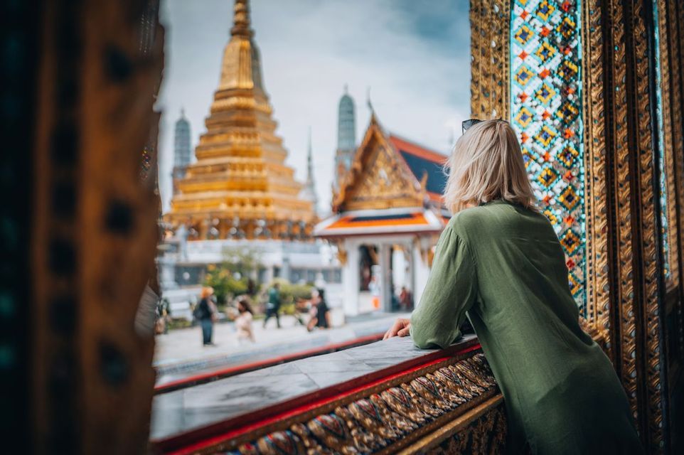 A blonde-haired person in a green shirt looks out from a decorated balcony at a golden Thai temple.