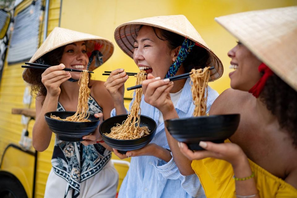 A WeRoad group trip of three women in conical hats laughing while eating noodles with chopsticks from black bowls.