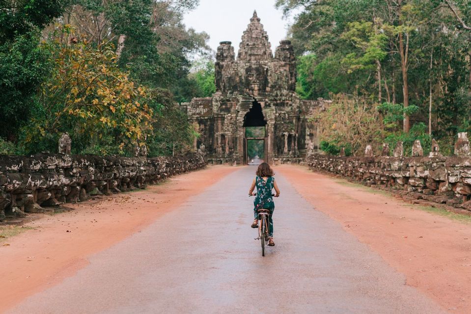 A woman rides a bicycle down a road towards an ancient stone temple gate flanked by statues and trees.