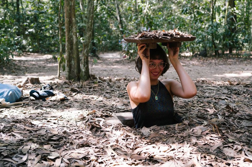 Una donna sorridente con gli occhiali emerge da una buca nel terreno ricoperta di foglie in una foresta, tenendo un coperchio sopra la testa.