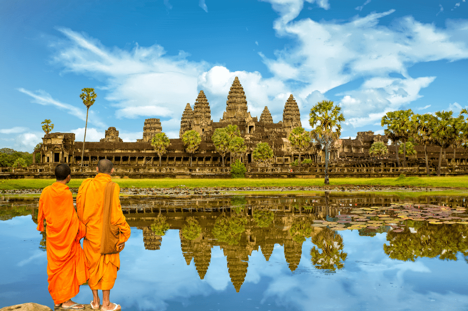 Two monks in orange robes stand by a lake, viewing an ancient temple complex reflected in the calm water under a blue sky.