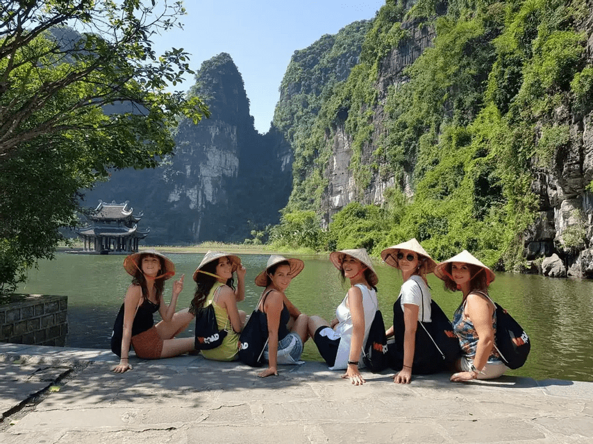 A WeRoad group trip of six women wearing conical hats, sitting on a stone ledge by a river with lush green karst mountains in the background.