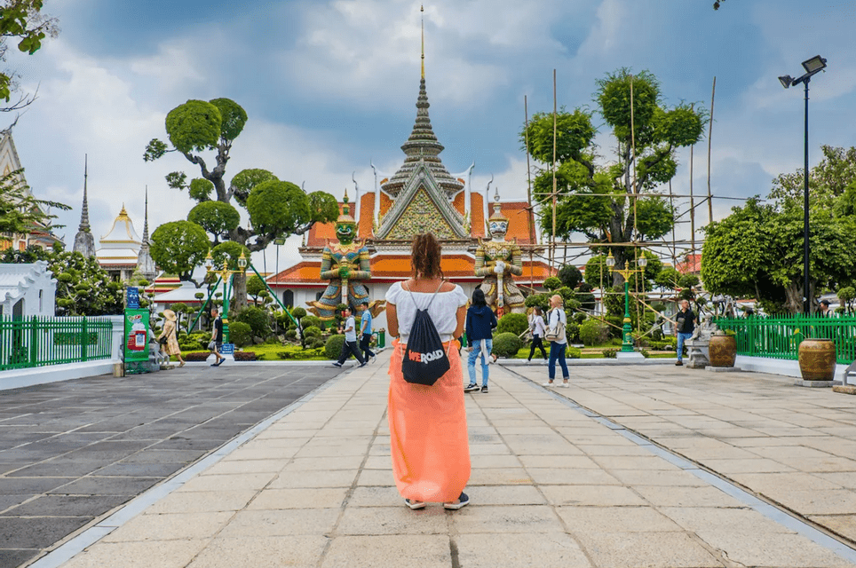 A woman viewed from behind, wearing a WeRoad backpack, looks towards an ornate temple with two giant guardian statues.