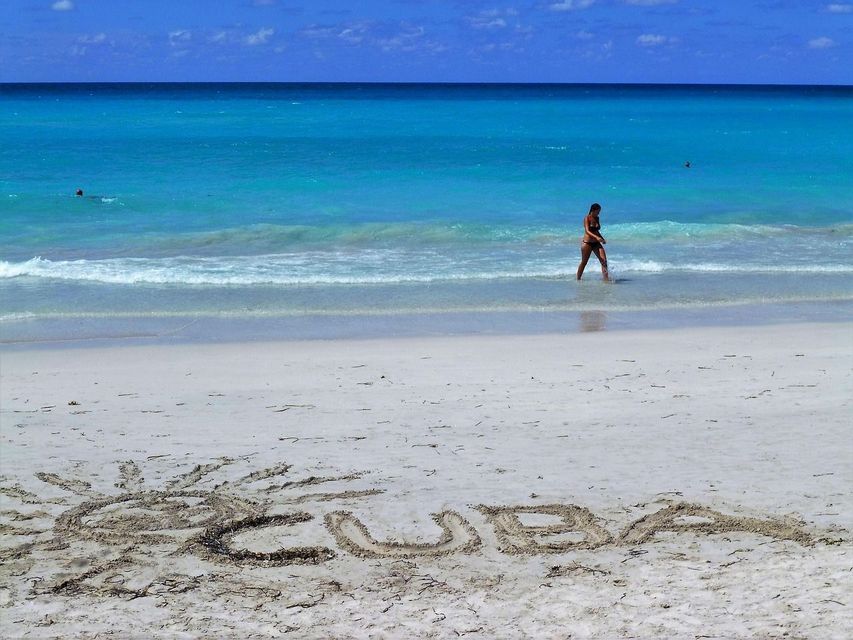 La palabra 'CUBA' y un sol están dibujados en la arena blanca de una playa mientras una persona camina en el océano turquesa.