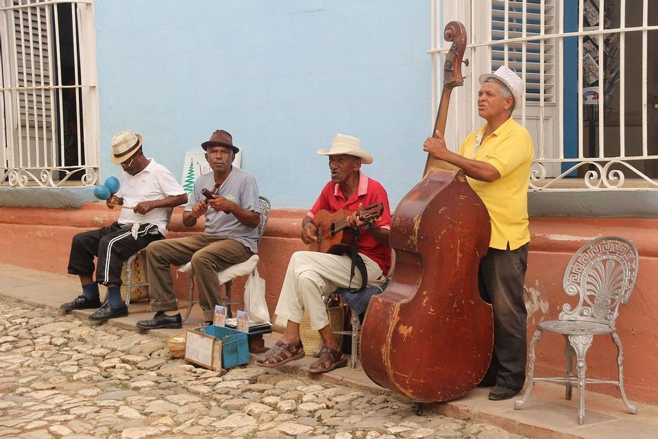 Quattro uomini che suonano un contrabbasso, una chitarra e strumenti a percussione su una strada di ciottoli accanto a un muro blu.