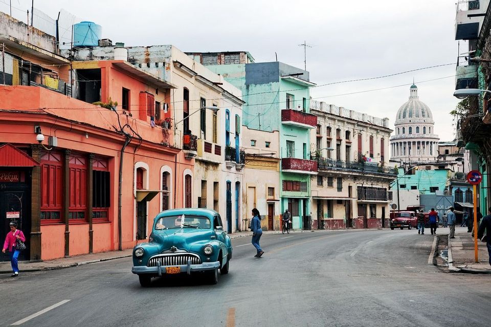 Una macchina blu d'epoca percorre una strada cittadina fiancheggiata da edifici colorati e logori, con un grande edificio a cupola sullo sfondo.