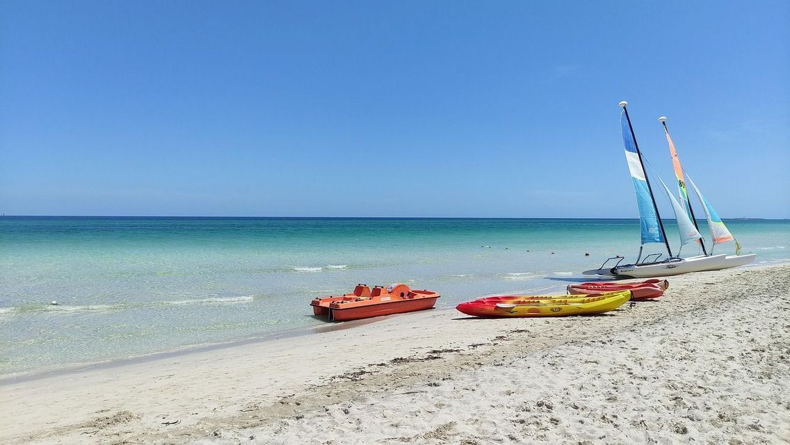 Catamarani, kayak e un pedalò sono adagiati sulla sabbia accanto all'acqua turchese sotto un cielo azzurro limpido.