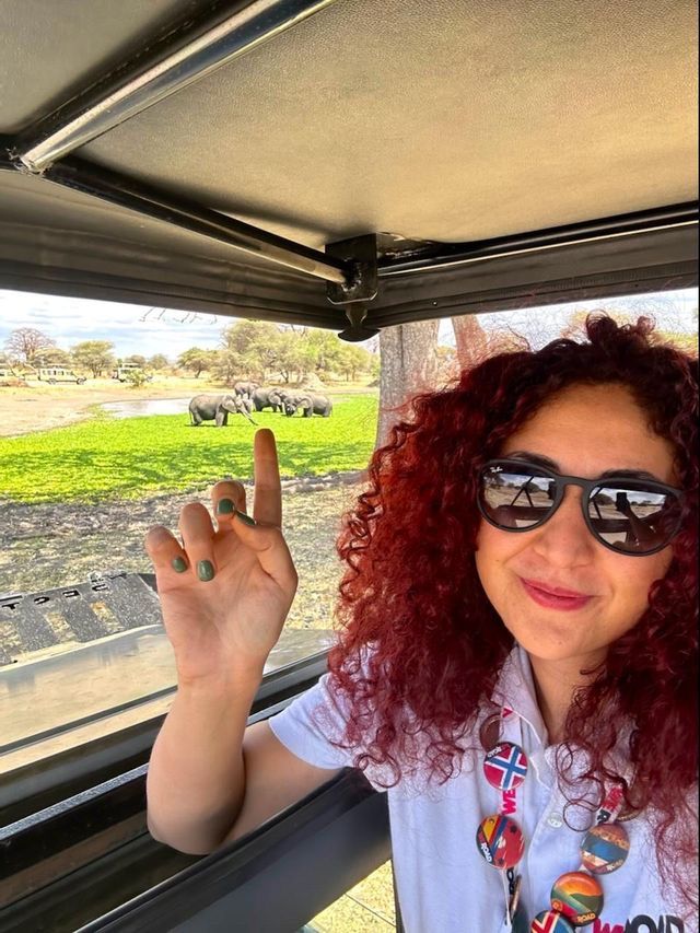Una donna con capelli rossi ricci e occhiali da sole si scatta un selfie all'interno di un veicolo safari, con una mandria di elefanti visibile sullo sfondo.