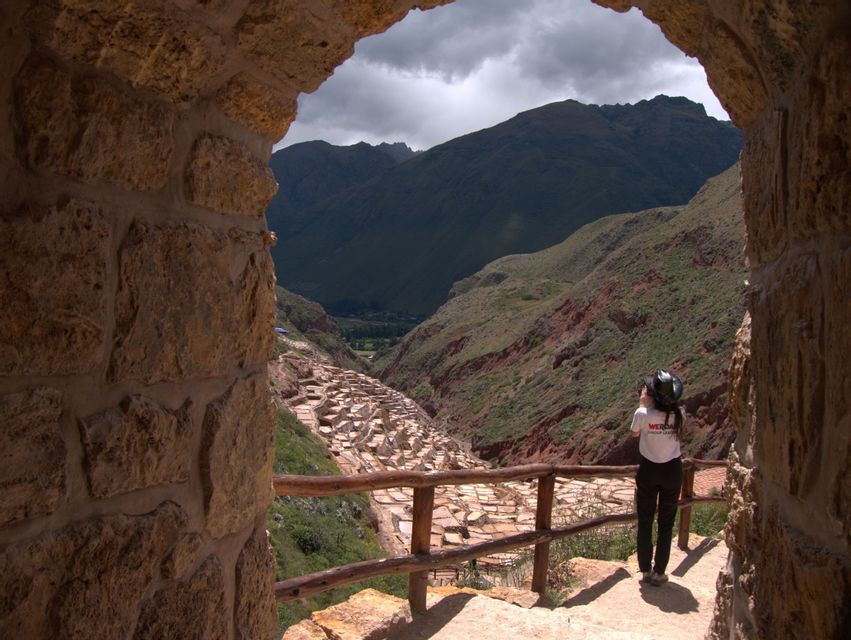 Una persona con una maglietta WeRoad si trova sotto un arco di pietra, ammirando le saline a terrazze in una valle di montagna.