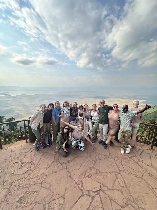 Un gruppo WeRoad in posa per una foto su un punto panoramico in pietra, con vista su una vasta valle e un lago sotto un cielo nuvoloso.