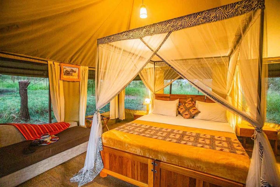 Un letto a baldacchino in legno con zanzariera e finiture stampa zebrata, all'interno di una tenda safari illuminata calorosamente, con vista su un'area esterna erbosa.