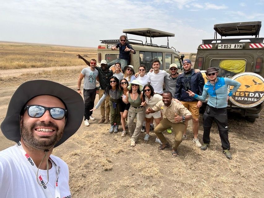 Un gruppo WeRoad si scatta un selfie sorridendo davanti a due jeep safari in una savana erbosa.