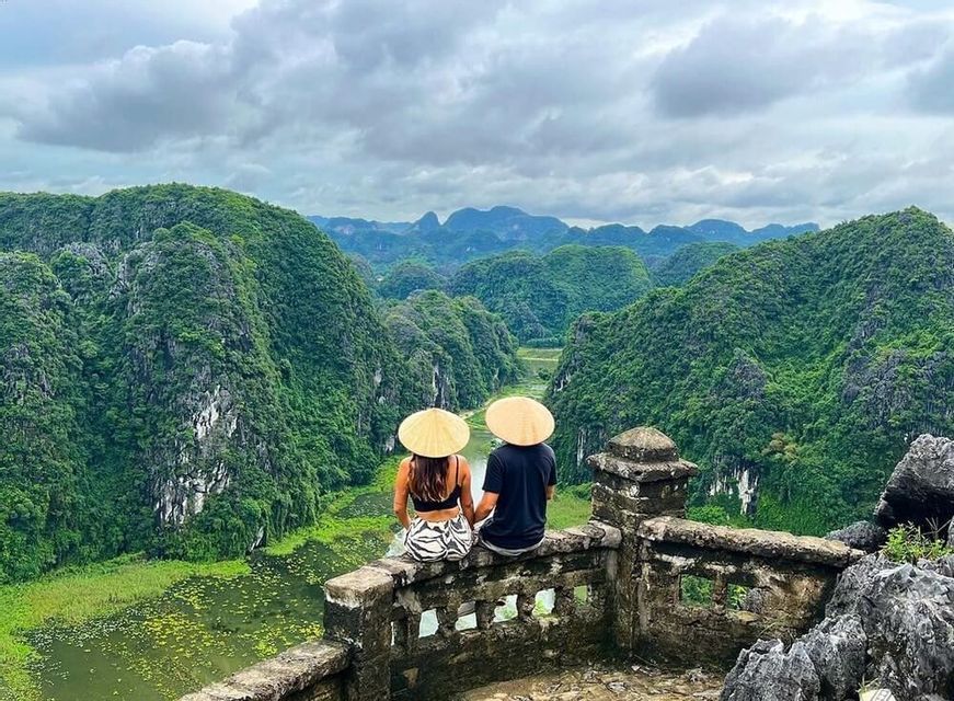A man and a woman wearing conical hats sit on a stone wall, looking out over a lush green river valley surrounded by mountains.