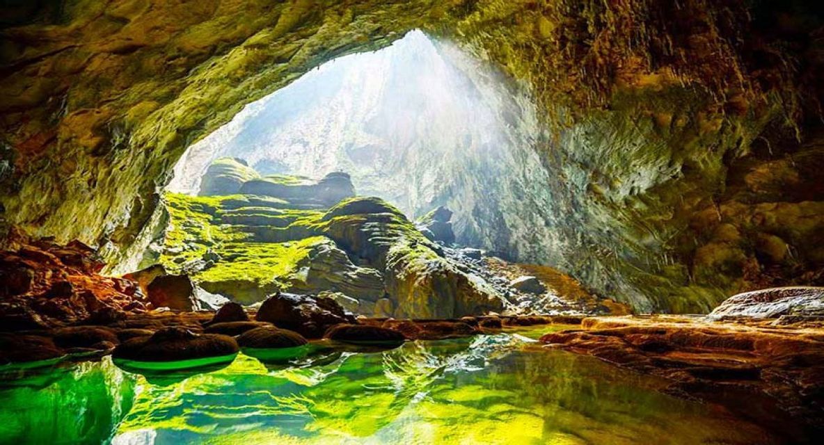 La luce del sole si riversa attraverso un'ampia apertura in una caverna, illuminando rocce coperte di muschio e una pozza d'acqua verde riflettente.