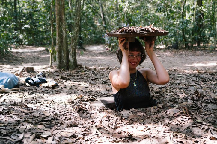 A smiling woman with glasses emerges from a hole covered with leaves in a forest, holding the leafy lid over her head.