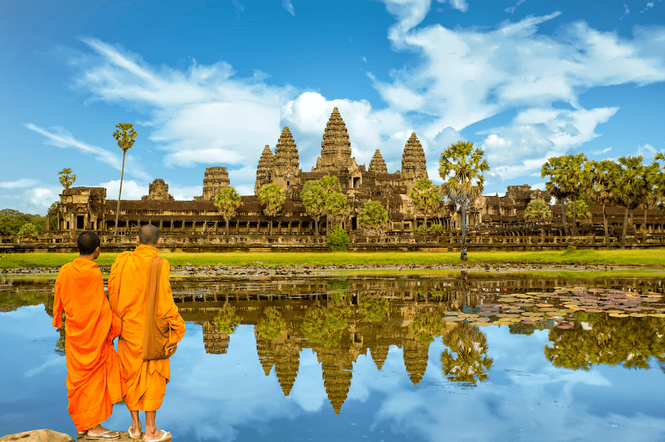 Two monks in orange robes stand by a lake, looking at an ancient stone temple reflected in the calm water.