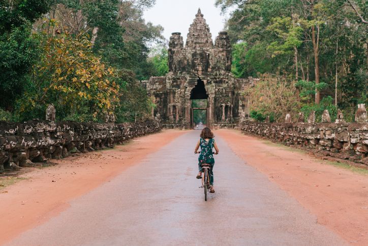A person seen from behind rides a bicycle on a road leading to a large, ancient stone gate flanked by jungle trees.