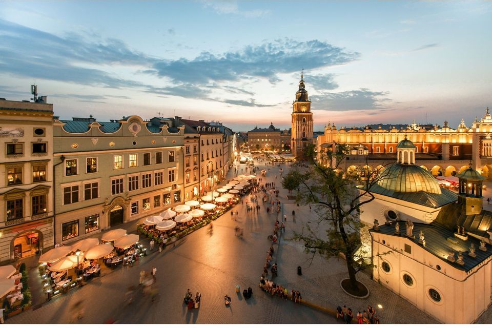 Ein erhöhter Blick auf einen historischen europäischen Stadtplatz in der Dämmerung, mit beleuchteten Gebäuden, Straßencafés und flanierenden Menschen.