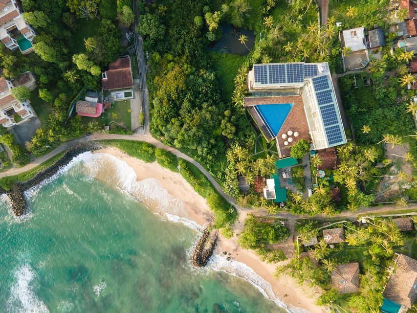 Luftaufnahme eines Hotels mit Solarpaneelen und dreieckigem Pool, direkt an einem Sandstrand mit türkisfarbenem Wasser.