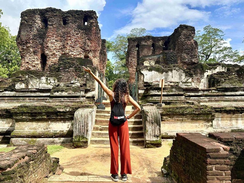 Une femme vue de dos, portant un sac à dos WeRoad, se tient devant d'anciennes ruines de briques couvertes de mousse, un bras levé.