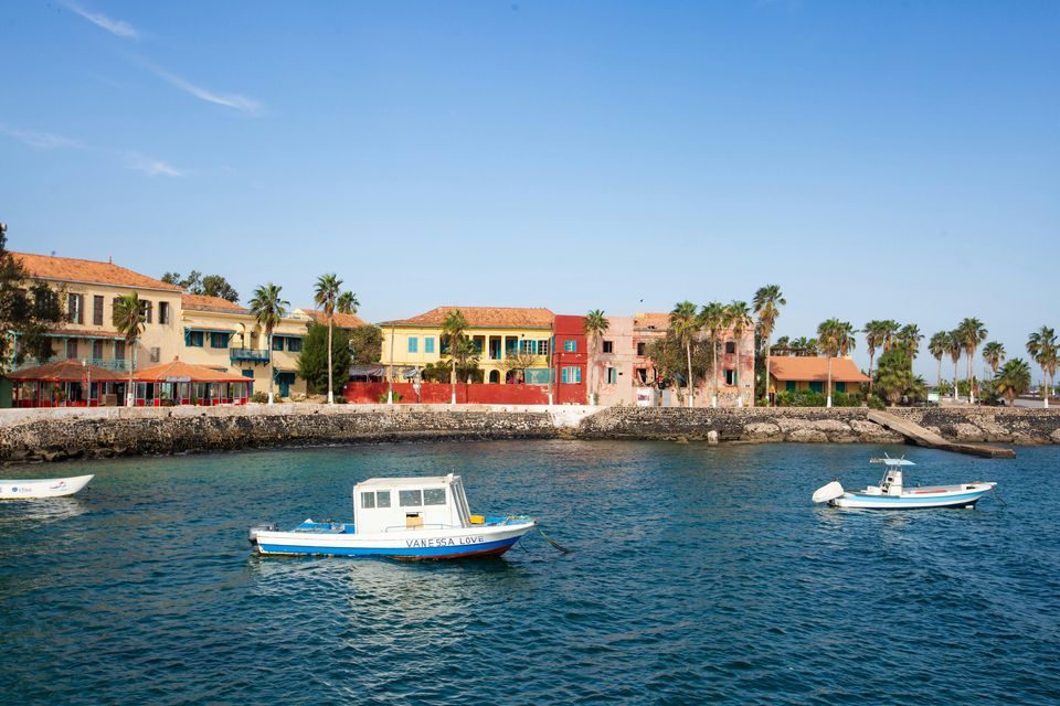 Pequeños barcos flotan en el agua frente a un colorido pueblo costero con palmeras bajo un cielo soleado.