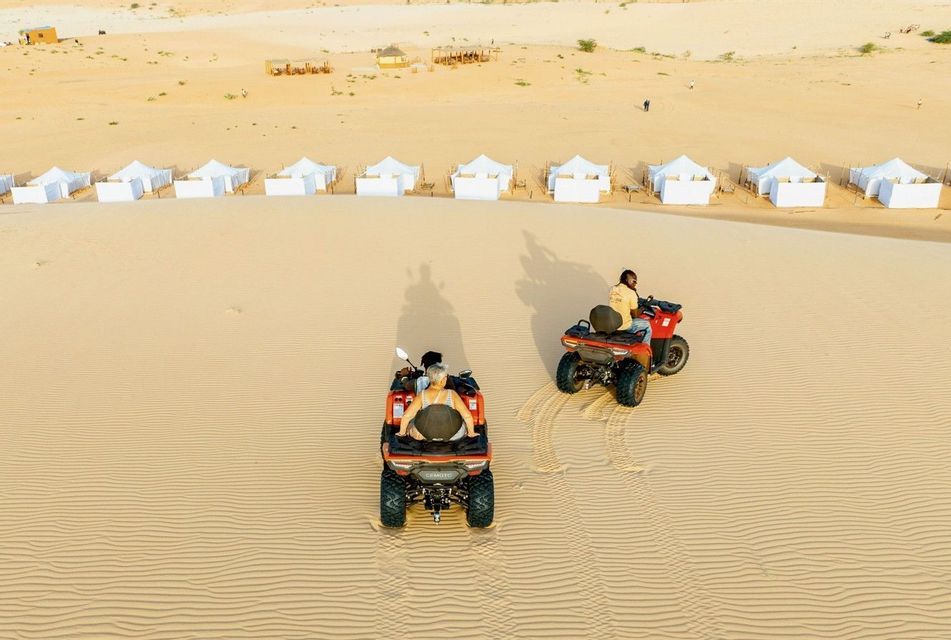 Vista aérea de dos personas de un viaje en grupo de WeRoad en quads por dunas de arena, con un campamento en el desierto visible al fondo.
