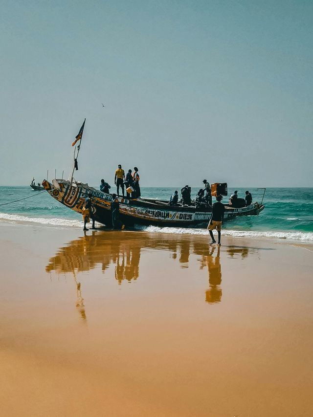 Un grupo de personas se reúne en y alrededor de una barca de madera tradicional en una playa de arena mojada junto al mar.