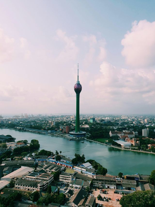 Ein hoher grüner Turm mit einer lotusförmigen Spitze steht inmitten einer Stadtlandschaft neben einem großen Gewässer.