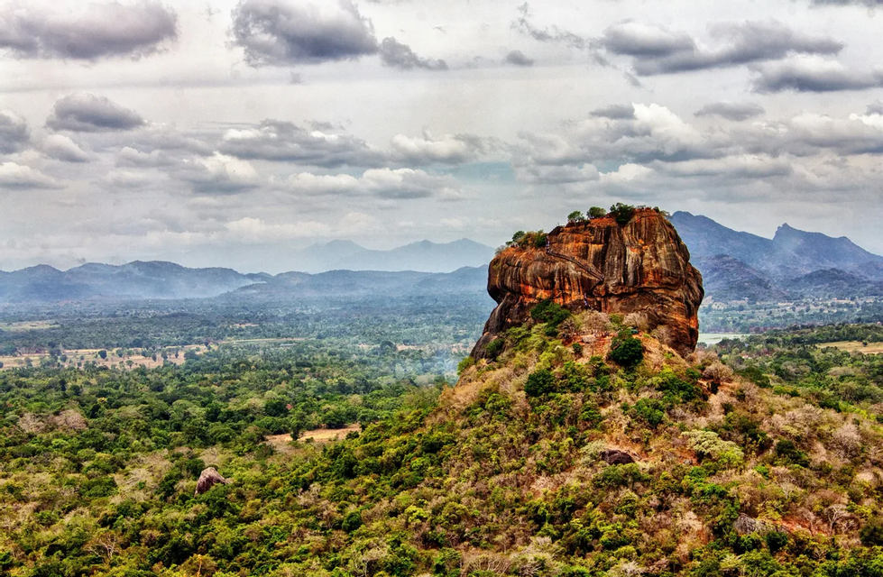 Un grand monolithe rocheux, brun-rougeâtre, s'élève au-dessus d'une vaste jungle verte et dense, avec des montagnes lointaines sous un ciel nuageux.