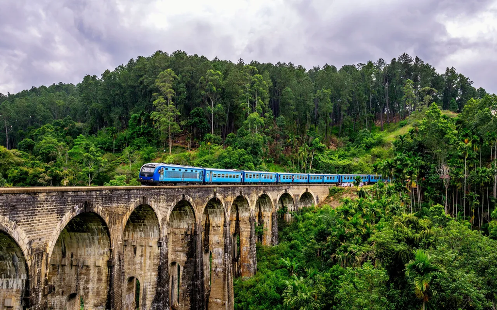 Ein blauer Zug fährt über eine hohe, mehrbogige Steinbrücke, umgeben von einem üppig grünen, tropischen Wald unter einem bewölkten Himmel.