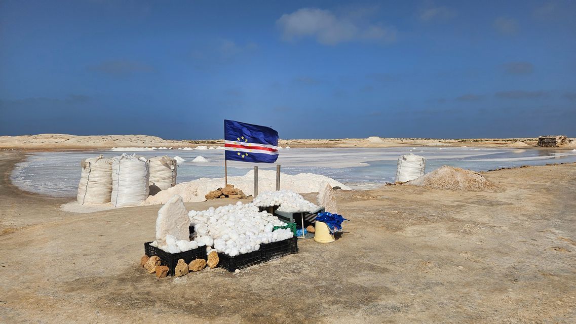 La bandera de Cabo Verde se alza entre pilas de sal cosechada en sacos y cajas en una salina bajo un cielo azul.