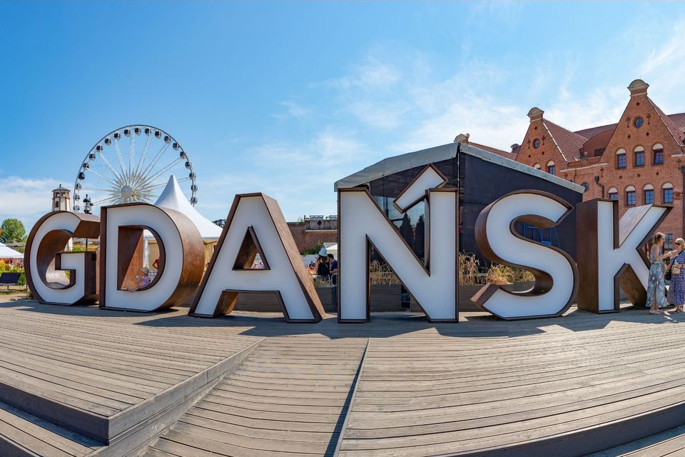 Ein großes, dreidimensionales Schild mit der Aufschrift 'GDAŃSK' auf einem Holzdeck, mit Riesenrad und Gebäuden im Hintergrund.