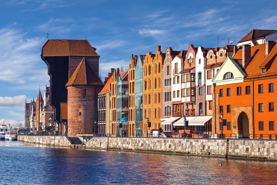 Eine Reihe farbenfroher Giebelhäuser und ein mittelalterlicher Backsteinkran säumen eine steinerne Uferpromenade und spiegeln sich unter blauem Himmel im Fluss.