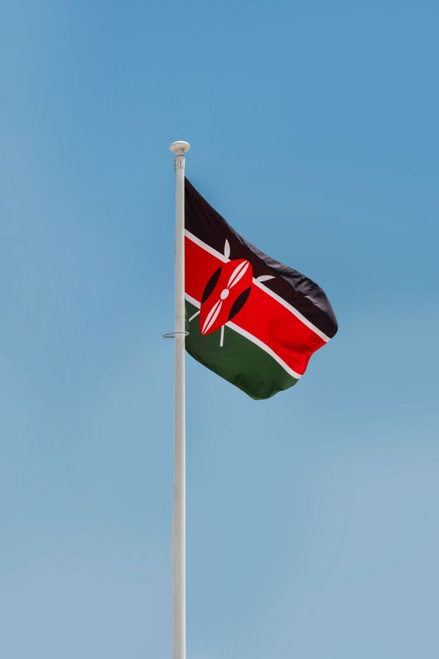 La bandera nacional de Kenia ondeando en un mástil blanco contra un cielo azul claro.
