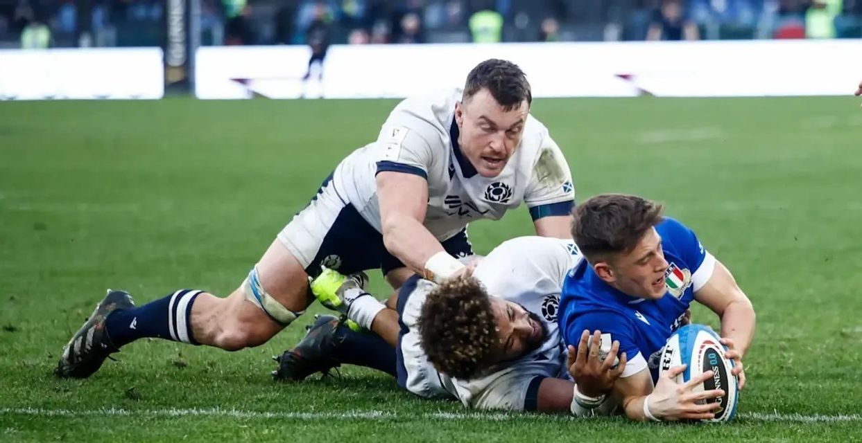Un giocatore di rugby in maglia blu viene placcato a terra da due avversari in maglia bianca mentre tiene la palla.