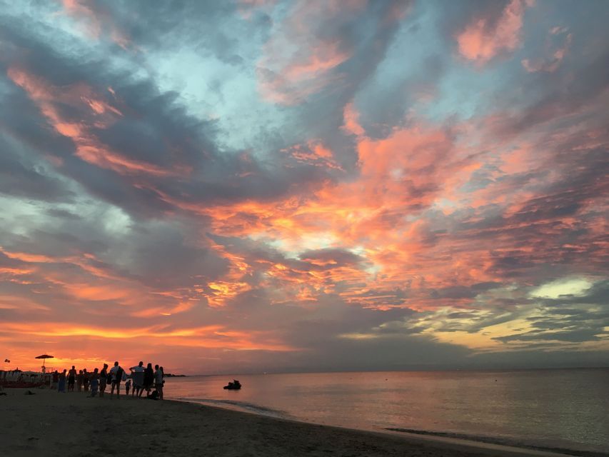Un gruppo WeRoad si trova su una spiaggia sabbiosa, ammirando un tramonto arancione e rosa vibrante sul mare calmo.