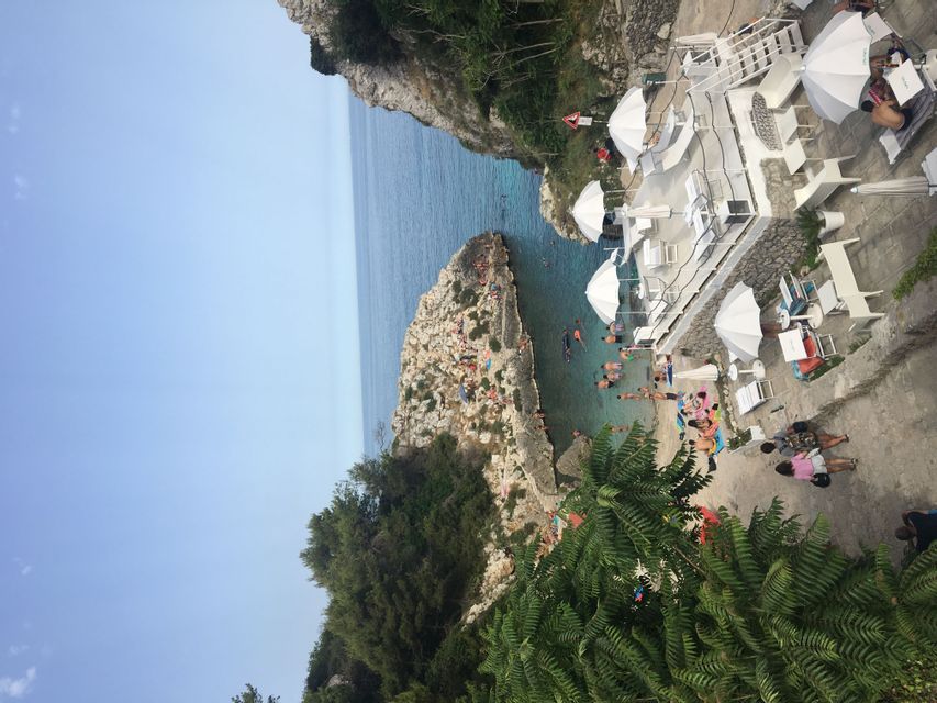 Vista dall'alto di una caletta rocciosa dove le persone nuotano e prendono il sole vicino a una terrazza su scogliera bianca con ombrelloni.