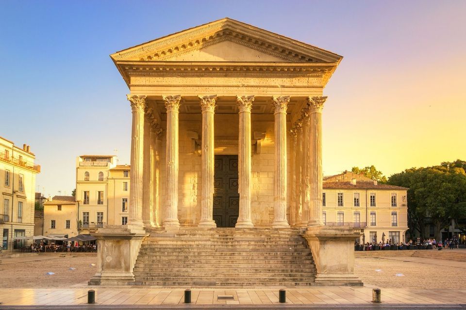 An ancient Roman-style temple with large columns and steps, standing in a paved square at sunset.