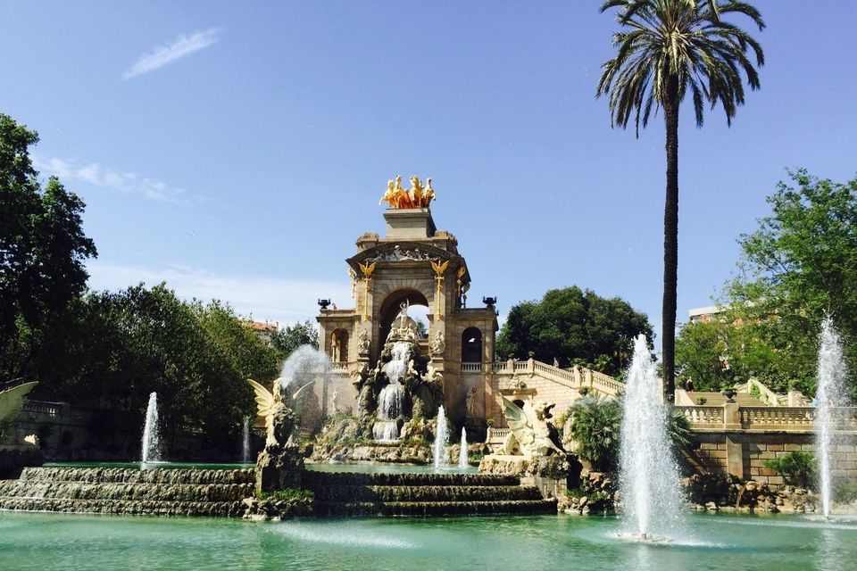 A large, ornate fountain with a golden sculpture sprays water into a pool, surrounded by lush trees and a tall palm tree under a blue sky.