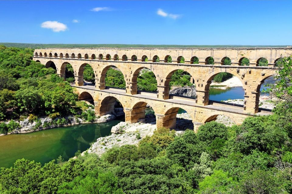 A three-tiered stone aqueduct with arches spans a river, surrounded by lush green forest under a bright blue sky.