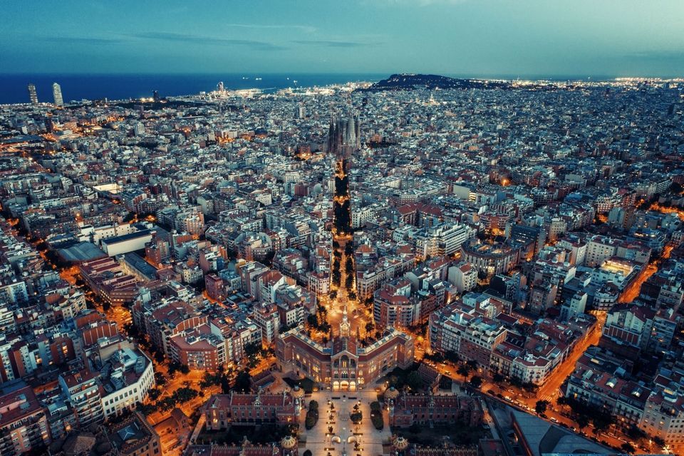 A high-angle view of a vast, illuminated cityscape at dusk, stretching towards the sea under a deep blue sky.