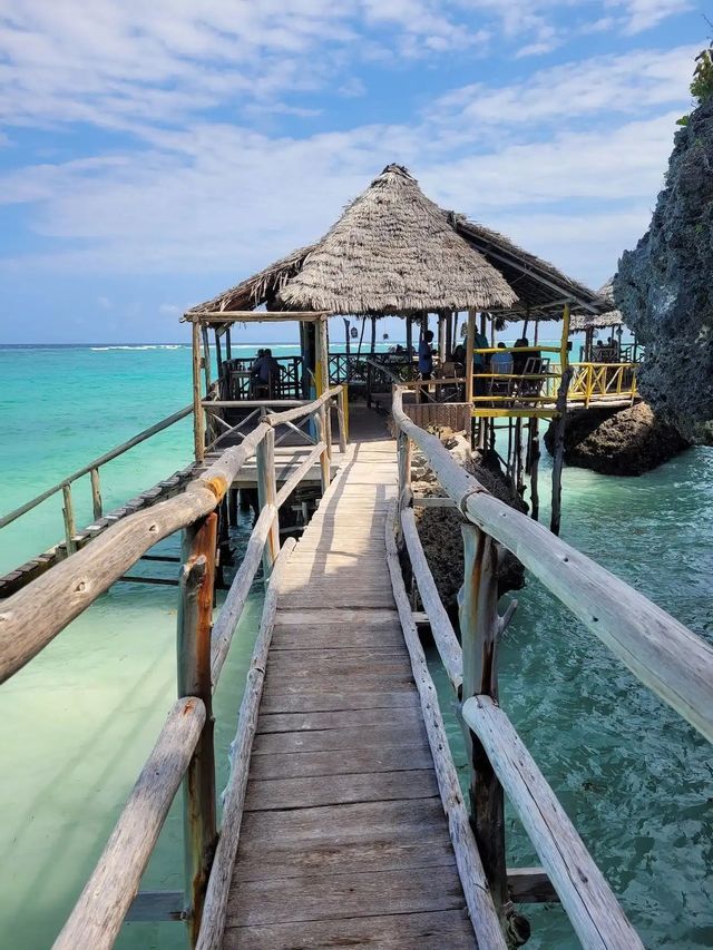 Una pasarela rústica de madera conduce a un restaurante con techo de paja construido sobre pilotes sobre aguas cristalinas de color turquesa.